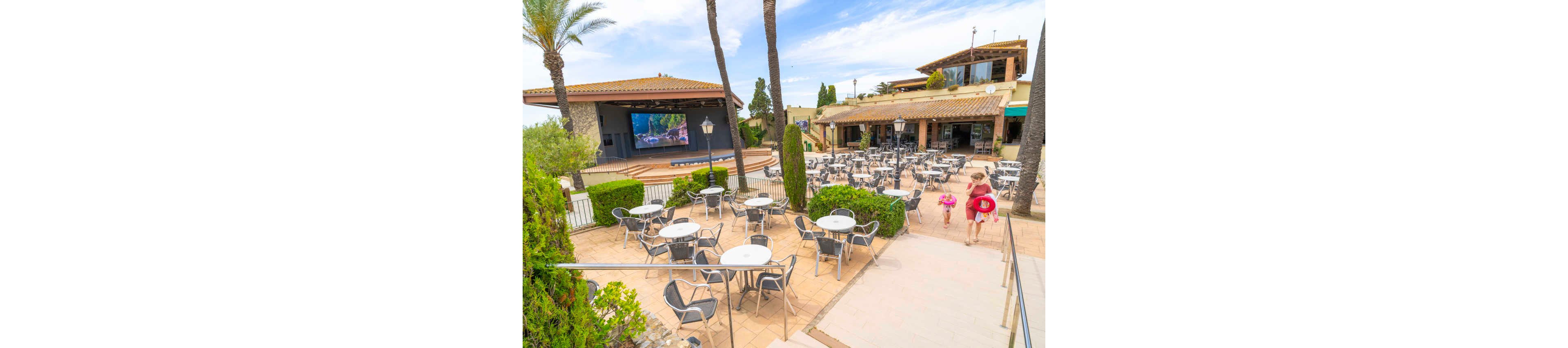 An open-air resort terrace features numerous empty metal tables and chairs arranged around a central area. A large outdoor stage with a big screen stands at the far end, framed by palm trees and Mediterranean-style buildings. Two children in swimsuits and float rings walk up the steps toward the seating area, suggesting a family-friendly holiday setting. The bright daylight and clear sky create a relaxed, summery atmosphere ideal for outdoor entertainment.