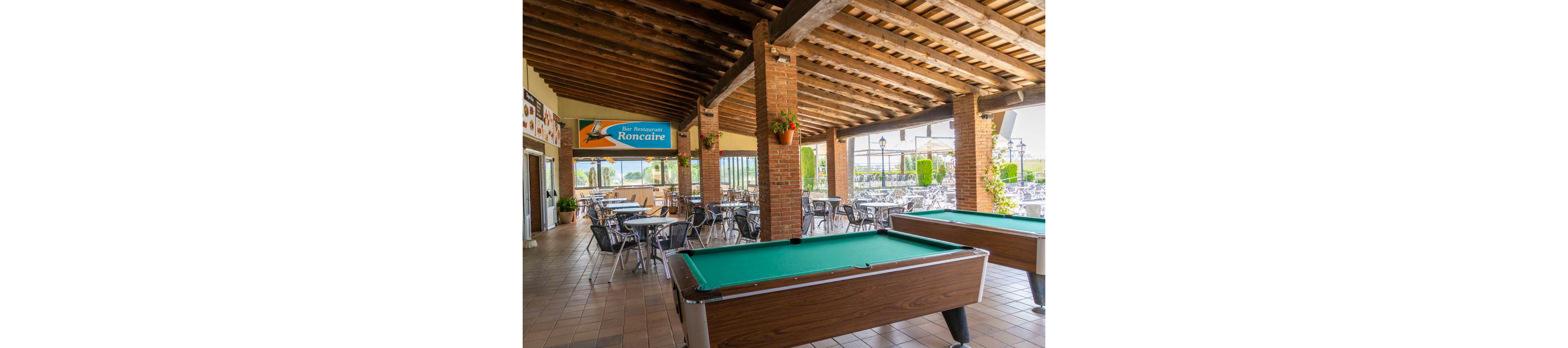 An open-air bar terrace features two green-felt pool tables in the foreground. Metal tables and chairs are arranged under a rustic wooden-beam roof supported by brick columns. Large windows and open sides reveal an outdoor seating area and distant landscape. Hanging plants and warm lighting create a relaxed, casual atmosphere. A sign reading "Bar Ristorante Roncaire" is visible above the seating area.