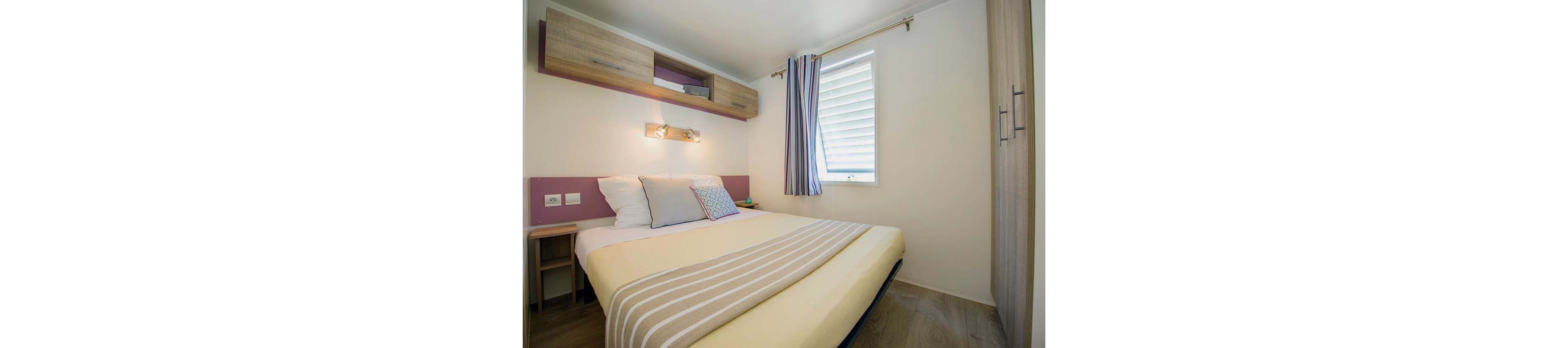 A compact modern bedroom features a neatly made double bed with a yellow cover and striped throw. The room includes built-in wooden cabinets above the headboard and a tall wardrobe on the right. A wall-mounted light fixture illuminates the bed, highlighting decorative pillows in soft pastel tones. A window with white blinds and striped curtains brings natural light into the simple, tidy space.