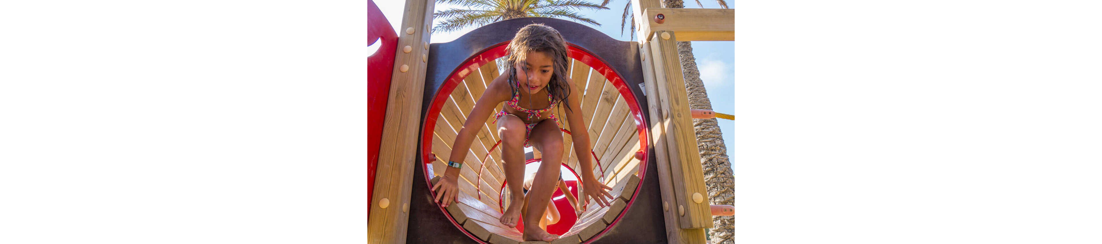 A young girl in a swimsuit crawls through a circular wooden tunnel on an outdoor playground. The structure features bright red accents and wooden beams, suggesting a beachside or park setting. Palm trees and a clear blue sky are visible in the background, creating a sunny, vacation-like atmosphere. The image captures an active, playful moment focused on movement and exploration.