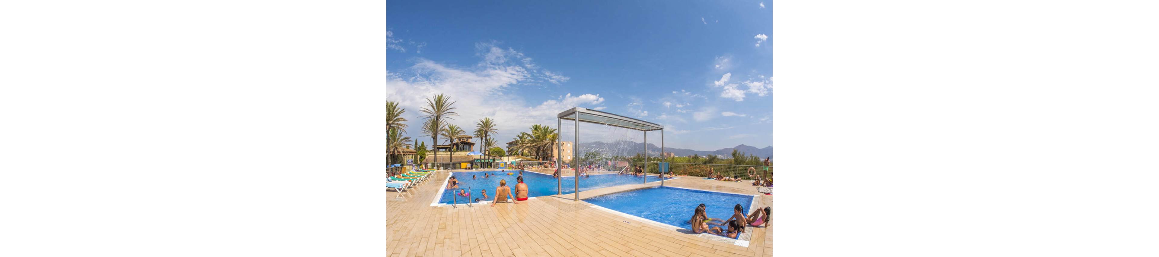 An outdoor resort swimming pool is filled with people relaxing and playing in the water. Families and children enjoy the pool under a clear blue sky with scattered clouds. Palm trees, lounge chairs, and distant buildings frame the scene, suggesting a warm vacation destination. A metal water feature pours a curtain of water into the pool, adding a playful element to the setting.