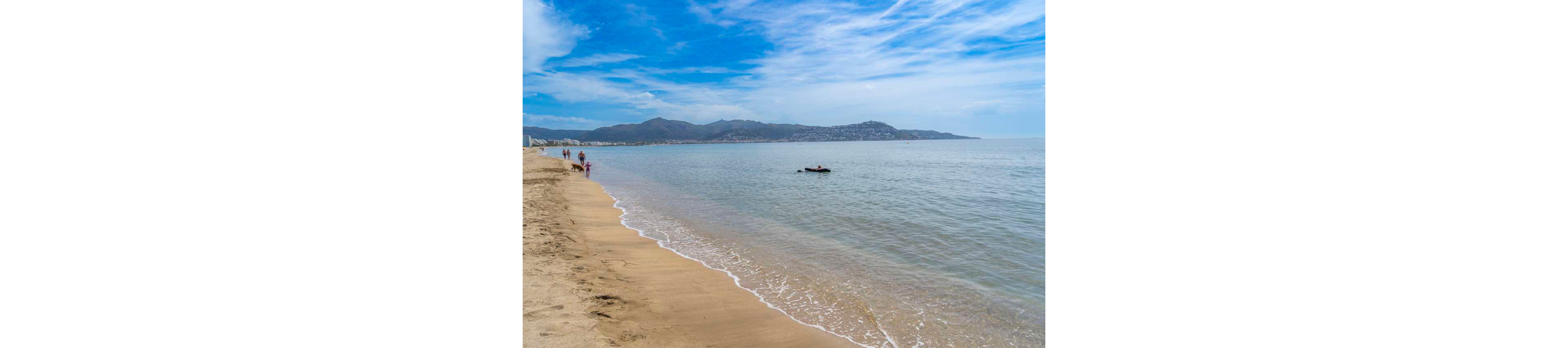 A wide sandy beach stretches along a calm shoreline under a partly cloudy blue sky. Gentle waves lap at the shore while a few people walk along the water’s edge in the distance. One person floats on the water further out, adding a sense of quiet leisure. Low mountains and a coastal town are visible across the bay, framing the tranquil seaside scene.