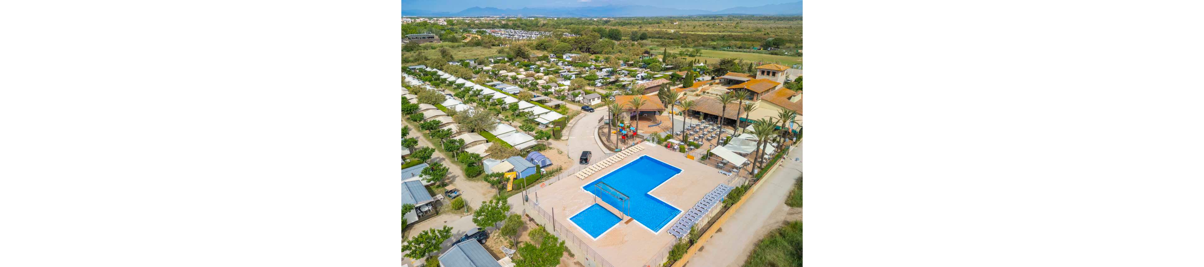 A large outdoor swimming pool is the central focus of this aerial view of a campsite resort. The pool area is surrounded by sun loungers and a fenced perimeter, with palm trees and landscaped greenery nearby. Rows of cabins, caravans, and camping plots extend into the distance, indicating a well-organized holiday park. In the background, open countryside and distant mountains frame the scene under clear skies.