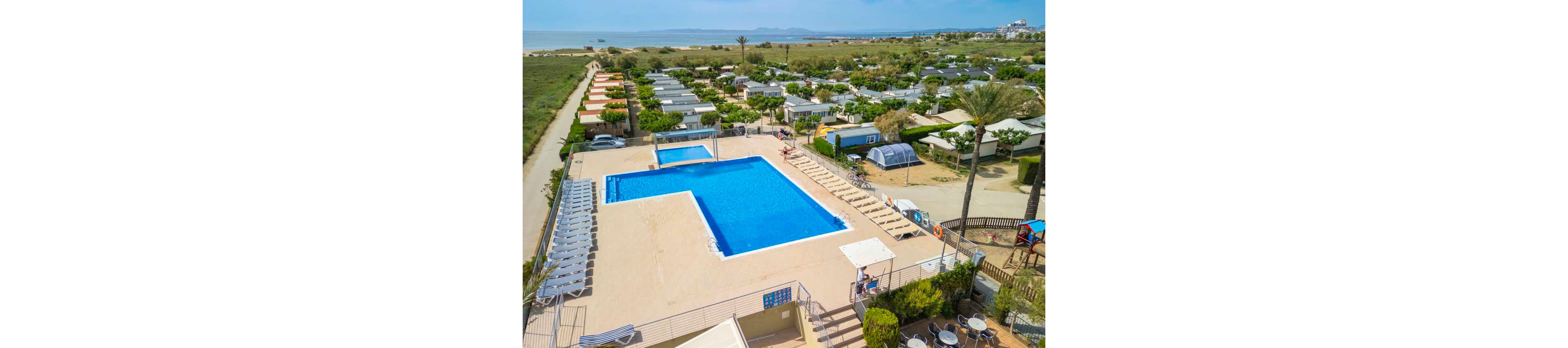 An elevated view shows a large rectangular swimming pool at the center of a campsite resort. Rows of sun loungers surround the pool area, with steps and railings leading down to the deck. Numerous mobile homes and bungalows extend into the distance, bordered by greenery and palm trees. Beyond the campsite, open fields and a coastline with calm water are visible under a clear sky. The scene suggests a relaxed holiday atmosphere in a coastal vacation park.