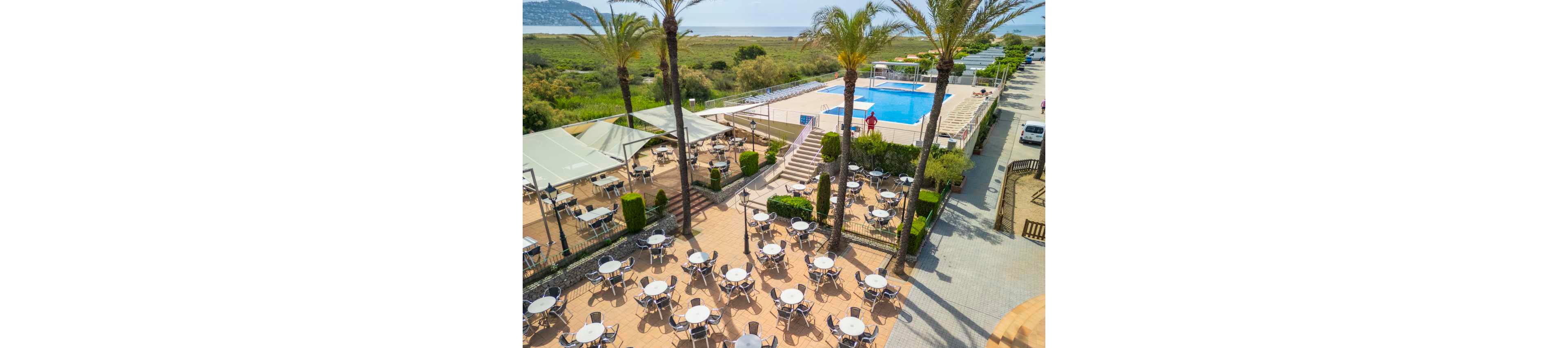 An elevated view shows a resort-style outdoor terrace filled with round tables and chairs arranged neatly on a tiled patio. A large rectangular swimming pool with clear blue water sits above the terrace, accessed by a central staircase. Palm trees and low hedges frame the seating area, while a lifeguard or staff member stands near the pool. In the background, natural greenery and distant hills suggest a coastal or countryside setting, and parked cars line a road beside the complex.