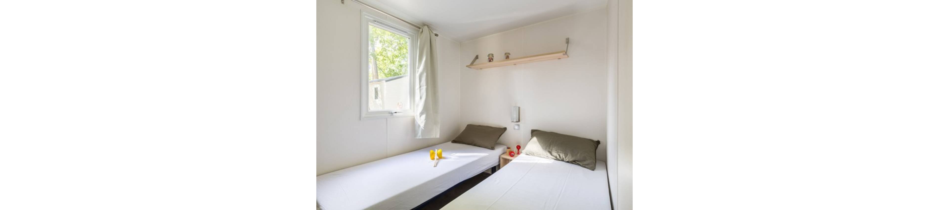 A small, bright bedroom features two simple twin beds with white linens and gray pillows. Natural light enters through a window with a light curtain, illuminating the clean white walls. A narrow wooden shelf above the beds holds a few decorative items, adding a subtle personal touch. On the bedside table between the beds, two yellow drinks and small red objects provide a pop of color in the otherwise minimalist space.