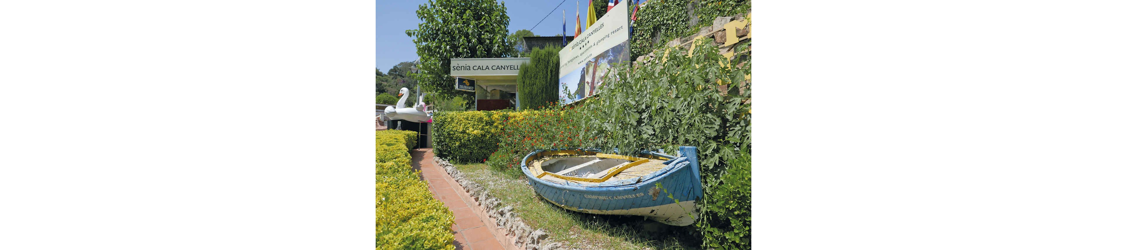 A weathered blue rowboat rests on a stone edge beside a lush garden at the entrance to Senia Cala Canyelles. Colorful flowers and dense green foliage surround the boat, creating a vibrant, summery atmosphere. In the background, a signboard and a large billboard promote the Senia Cala Canyelles resort, while flags rise above the greenery. A white swan-shaped structure is visible further along the path, adding a playful touch to the scene.