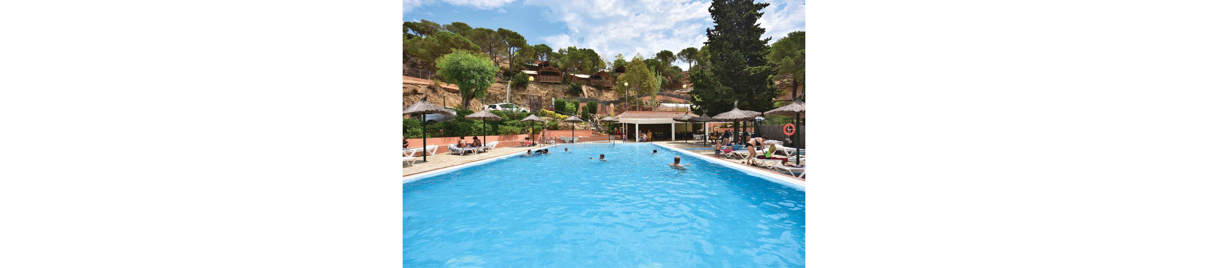 A large outdoor swimming pool is surrounded by sun loungers and straw parasols at a hillside resort. Guests relax on chairs and swim in the clear blue water under a bright sky with scattered clouds. Behind the pool, terraced paths, trees, and small cabins rise up the slope, creating a secluded holiday atmosphere. A poolside bar or pavilion sits at one end, adding to the leisurely vacation setting.