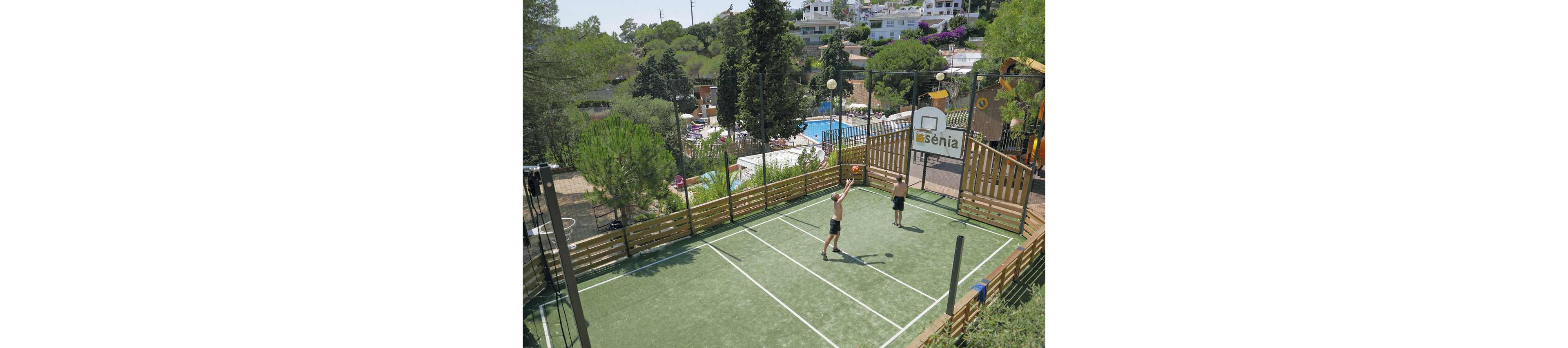 Two teenage boys play basketball on a small fenced outdoor court with a green surface. One player prepares to shoot while the other watches near the hoop. The court is surrounded by wooden barriers and tall netting, with trees and a swimming pool area visible in the background. A sign with the word "sènia" appears above the basketball hoop, suggesting a recreational or holiday complex setting.