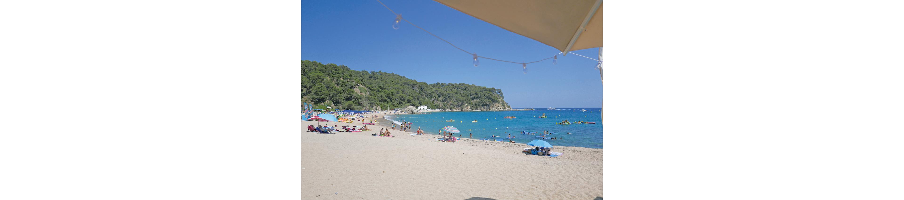 A wide sandy beach is filled with people relaxing under umbrellas and sunbathing near the shoreline. Swimmers and paddleboarders enjoy the clear blue sea, with inflatable floats and small watercraft scattered across the water. Green hills covered in trees frame the bay in the background under a cloudless sky. A large beach umbrella and string lights are visible at the top of the image, adding a casual holiday atmosphere.