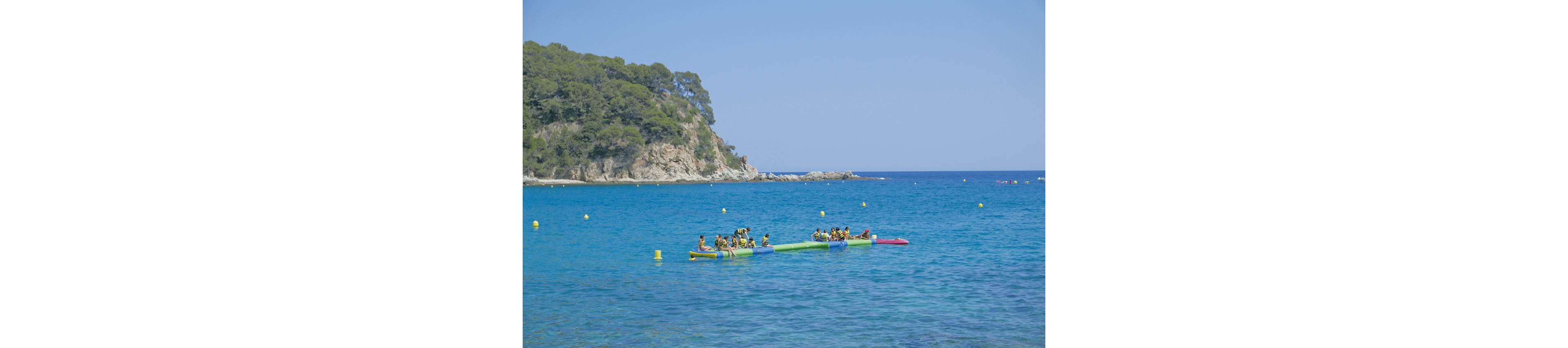 A group of people play on colorful inflatable structures floating on clear blue sea water. The scene is set near a rocky, tree-covered coastline under a bright, cloudless sky. Yellow buoys dot the water, marking the swimming or activity area. The overall atmosphere is summery, relaxed, and playful, suggesting a seaside vacation or beach resort.