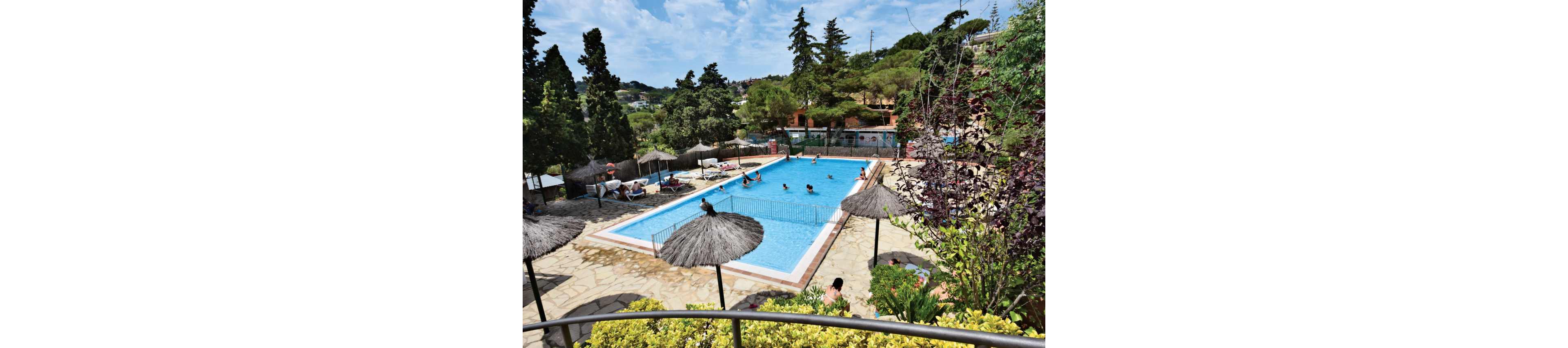An outdoor rectangular swimming pool is surrounded by sun loungers and straw parasols at a hillside resort. Several people are swimming and playing in the water while others relax on chairs around the pool. Lush greenery, trees, and landscaped gardens frame the pool area, creating a tranquil holiday atmosphere. The scene is captured on a bright day with blue sky and scattered clouds overhead.