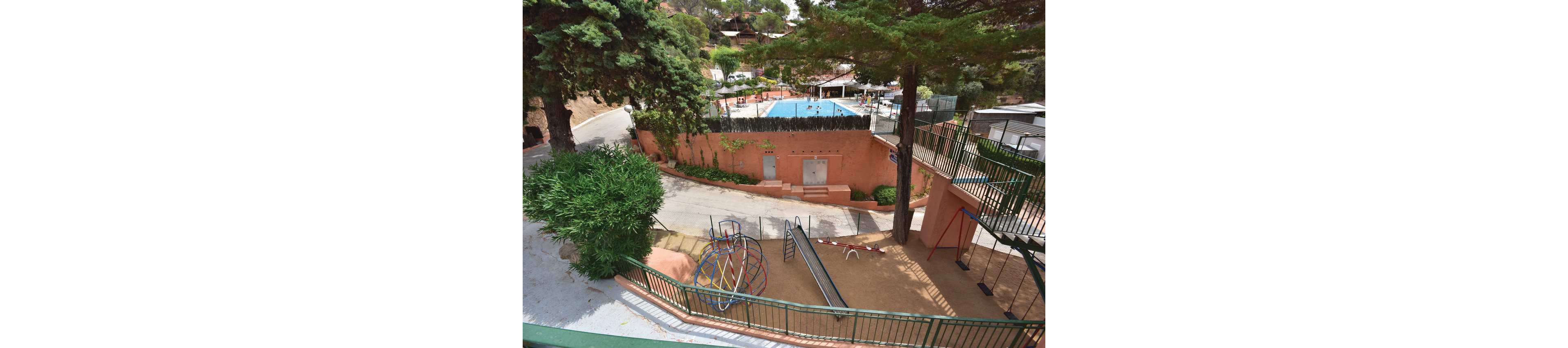 An outdoor recreation area features a small playground with slides, swings, and a climbing dome on a sandy surface. The playground is enclosed by green metal railings and shaded by tall trees. Beyond a peach-colored wall, a rectangular swimming pool with blue water is visible, surrounded by sun loungers and umbrellas. The scene suggests a family-friendly leisure space in a warm, sunny climate.