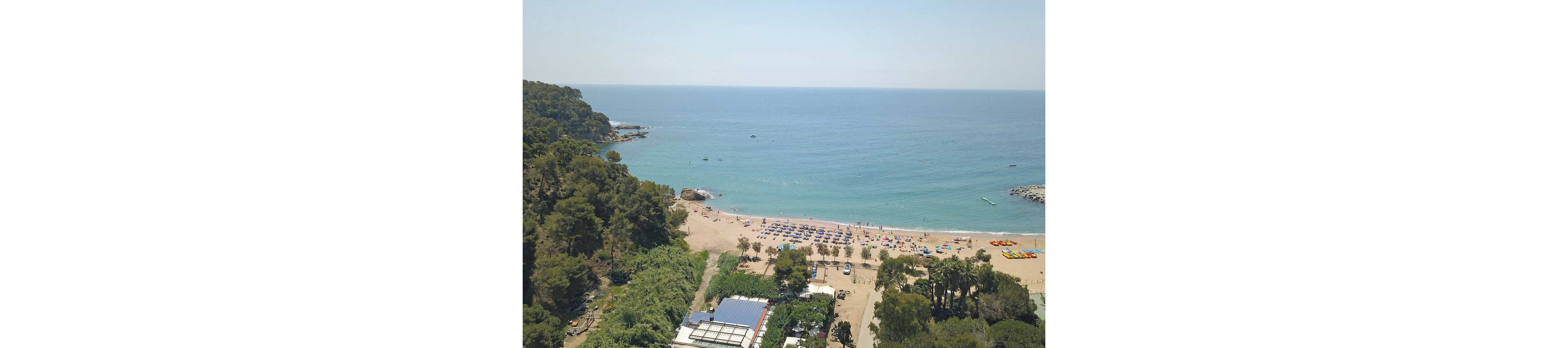 A wide sandy beach is seen from above, lined with rows of umbrellas and sun loungers near the shoreline. The calm blue sea stretches to the horizon with a few small boats and swimmers visible in the water. Dense green trees and vegetation frame the beach on both sides, with some buildings and structures nestled among the foliage. The scene suggests a popular summer holiday destination with a relaxed coastal atmosphere.