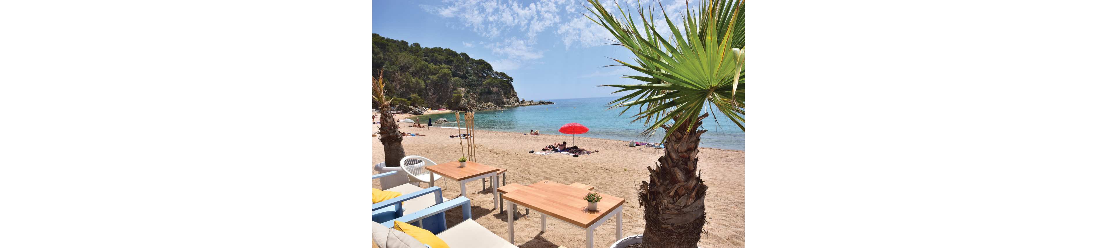 A relaxed beachside cafe with wooden tables and colorful chairs overlooks a sandy shoreline. Palm trees frame the foreground while people sunbathe and lounge near the water. A bright red umbrella stands out on the sand against the calm blue sea. Green hills and scattered clouds complete the peaceful coastal scene.