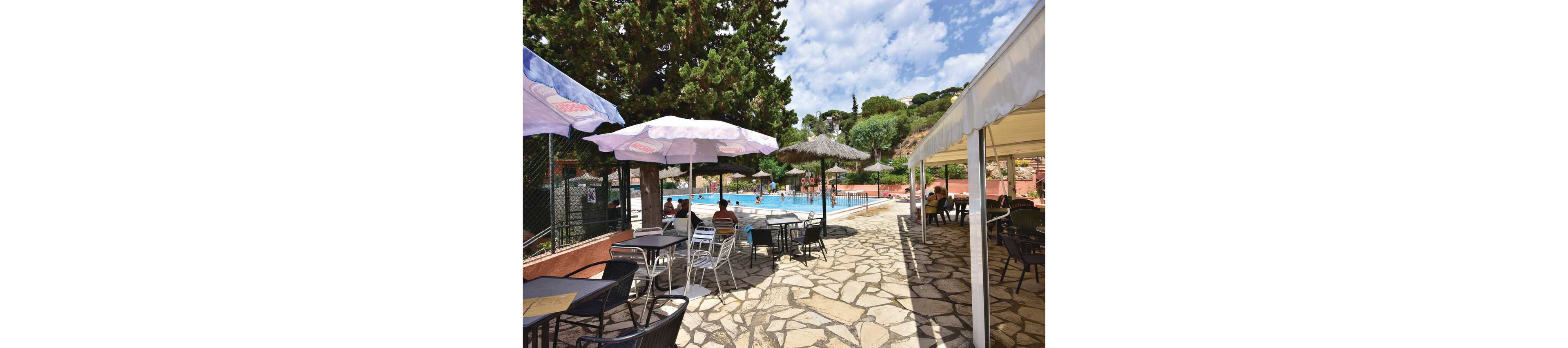 An outdoor swimming pool is surrounded by a stone patio and shaded seating area. Tables and chairs sit under large umbrellas and a canopy, creating a relaxed poolside café atmosphere. People are swimming and lounging in the background, enjoying a sunny day. Tall trees and green hills frame the scene under a partly cloudy sky.