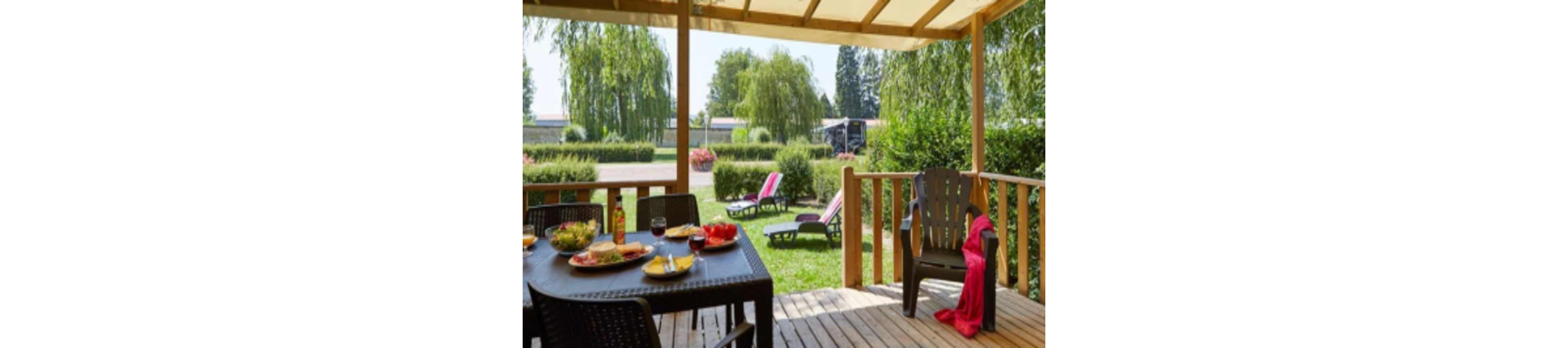 A wooden terrace overlooks a lush green garden with several lounge chairs arranged on the grass. On the patio table, plates of food, salad bowls, and glasses of red drink suggest an outdoor meal. A plastic armchair with a bright red towel draped over it adds a pop of color. In the background, trees, hedges, and a parked vehicle create a relaxed vacation or campsite atmosphere under a clear sky.