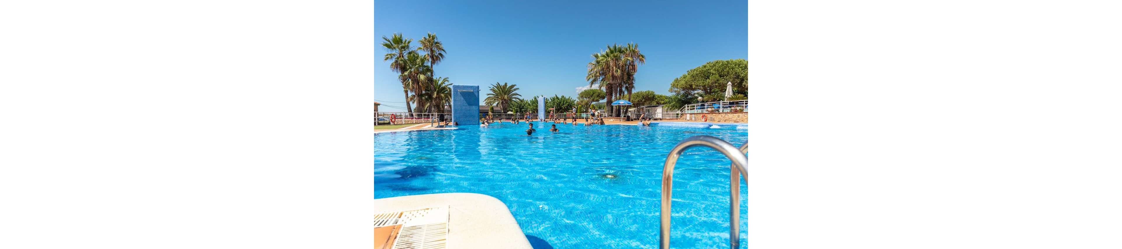 A large outdoor swimming pool sparkles under a clear blue sky. Several people are swimming and playing in the water, enjoying a warm sunny day. Palm trees and green foliage surround the pool area, giving it a tropical resort atmosphere. Poolside loungers, umbrellas, and railings frame the scene, suggesting a relaxed vacation setting.
