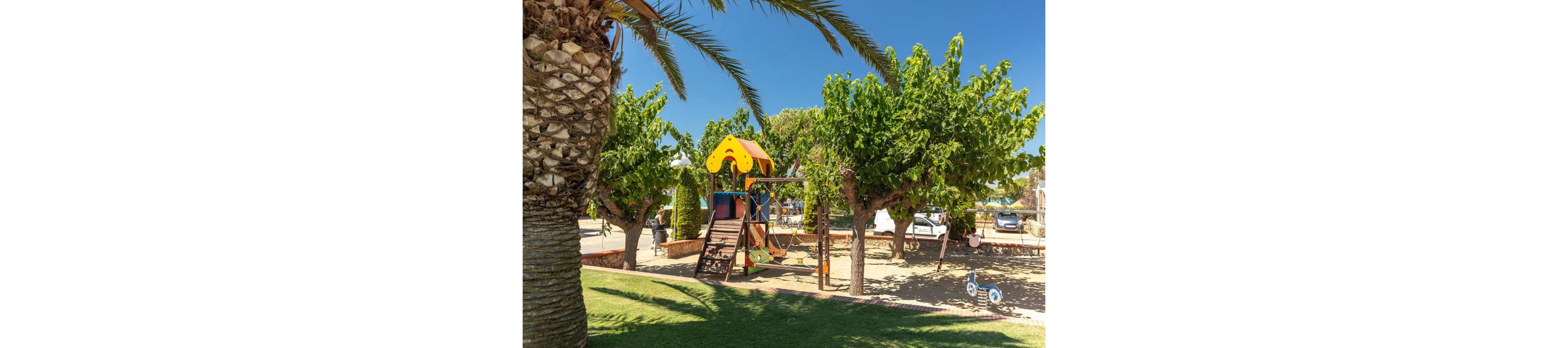 An outdoor playground with a colorful slide and swings sits beneath leafy trees on a bright sunny day. The play structure stands on sandy ground, surrounded by grass and a few palm trees. In the background, parked cars and low buildings suggest a park or campground setting. The clear blue sky and strong sunlight create a cheerful, family-friendly atmosphere.