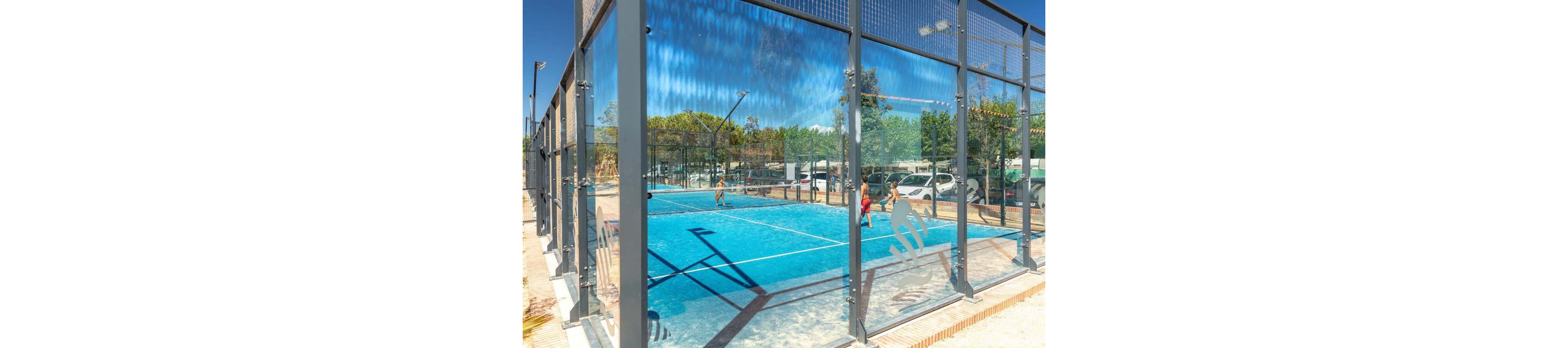 An outdoor padel court with bright blue flooring is enclosed by tall glass and metal walls. Several people are playing a game under a clear blue sky, surrounded by trees and parked cars in the background. Sunlight reflects off the glass panels, creating dynamic reflections of the court and players. A stylized padel player graphic is visible on the glass near the foreground.