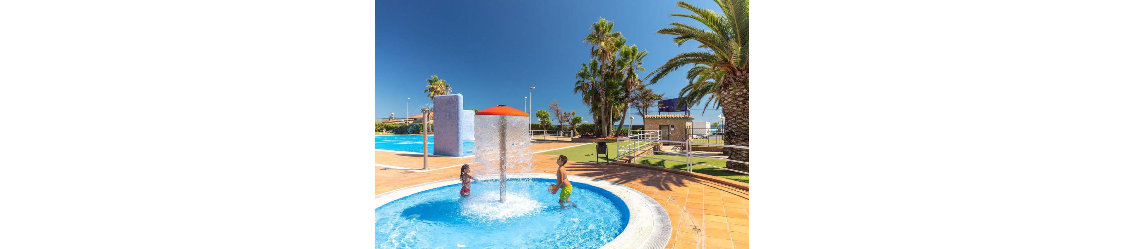 Two children play in a shallow circular splash pool with a central mushroom-style water fountain. The scene is set in an outdoor resort pool area with bright blue water and tiled pool decks. Palm trees, green lawns, and distant buildings frame the sunny, cloudless sky. The overall atmosphere is warm, playful, and summery, suggesting a family-friendly vacation destination.