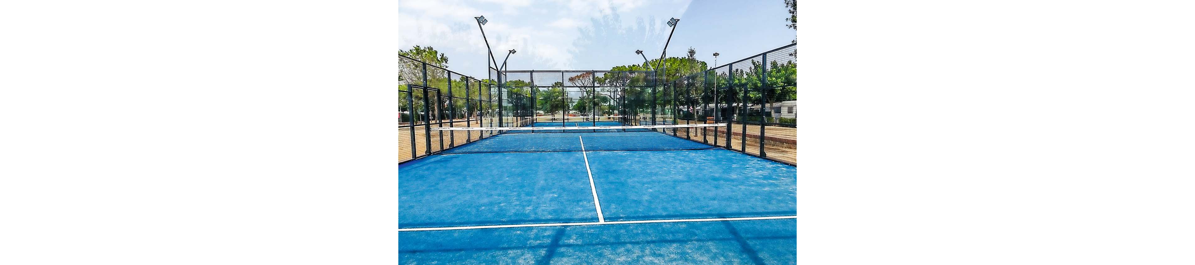 An empty blue padel court is shown from the baseline, framed by tall metal fencing and glass walls. The court is located outdoors in a sports facility surrounded by trees and greenery. White court lines and a central net divide the playing area under a bright, partly cloudy sky. No players are present, emphasizing the clean surface and recreational setting.