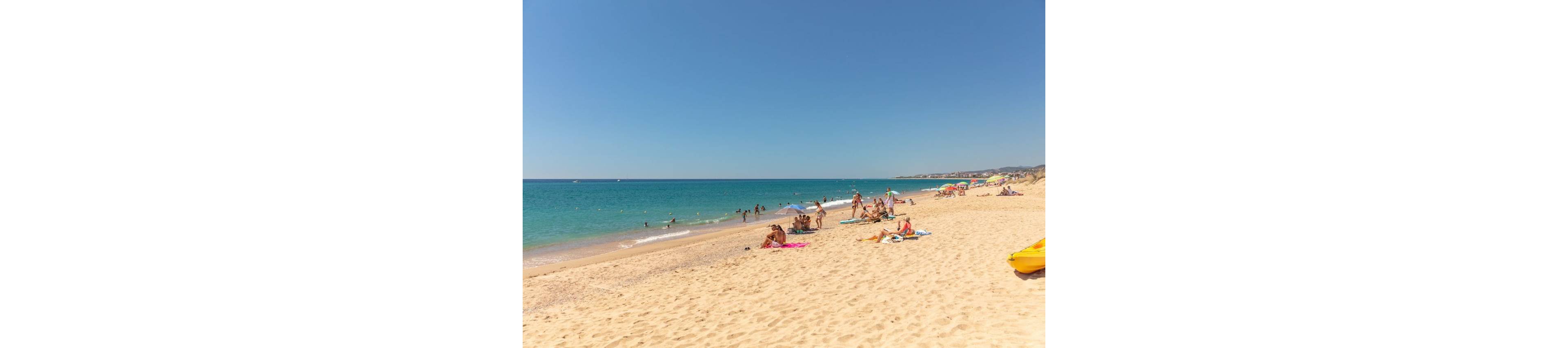 A wide sandy beach is filled with people relaxing and sunbathing under a clear blue sky. Swimmers and waders enjoy the calm turquoise sea near the shoreline. Towels, umbrellas, and beach gear are scattered across the warm sand. The bright daylight and clear horizon create a relaxed summer vacation atmosphere.