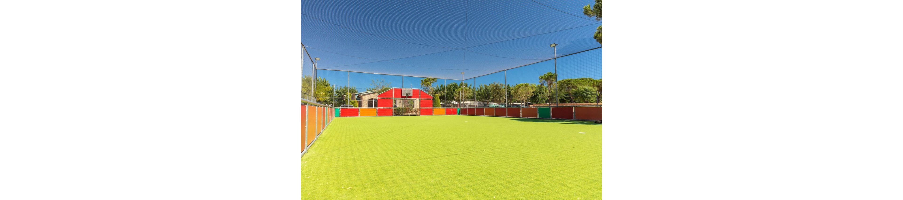 An empty artificial turf sports field is surrounded by brightly colored panels in an outdoor recreational area. Netting stretches overhead, enclosing the entire pitch beneath a clear blue sky. Trees and small buildings are visible in the background, suggesting a park or holiday resort setting. The vibrant colors and sunny weather create a cheerful, family-friendly atmosphere.
