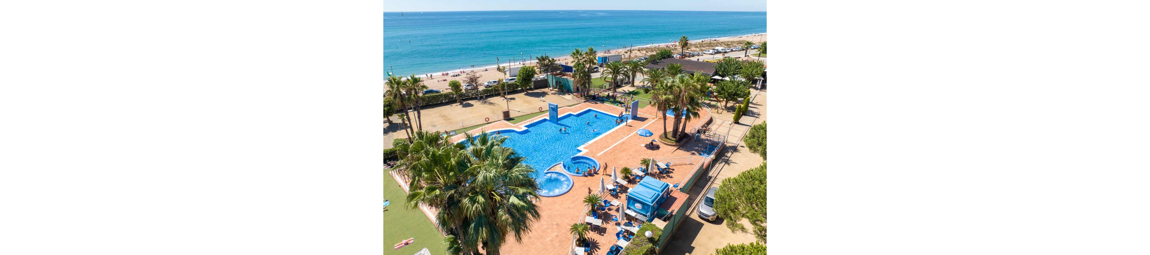 A large outdoor swimming pool complex sits beside a sandy seaside beach. People relax on sun loungers and swim in the main pool and a smaller circular pool. Tall palm trees, umbrellas, and deck chairs create a tropical holiday atmosphere. The bright blue sea and clear sky stretch into the distance, emphasizing a sunny coastal vacation setting.