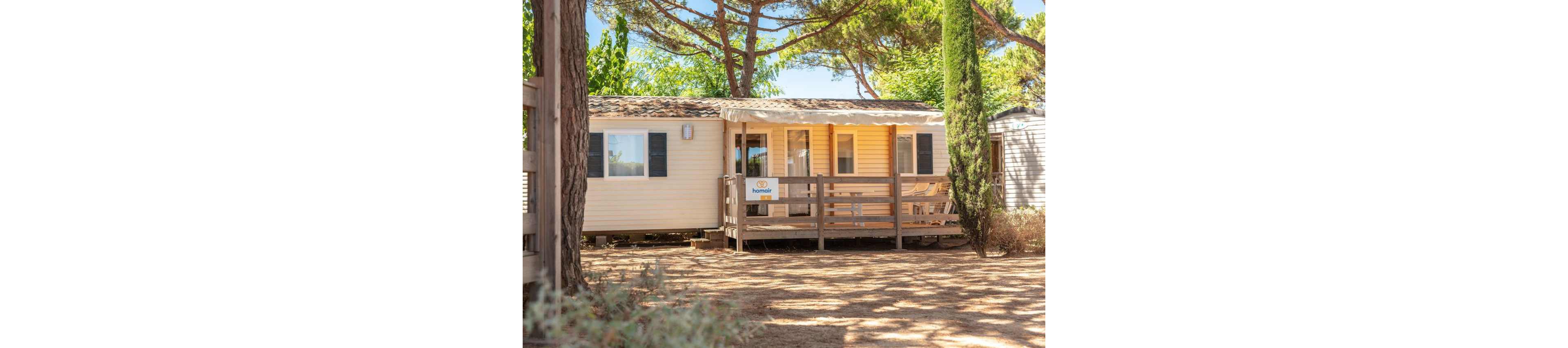 A light-colored mobile home with a shaded wooden terrace stands among tall trees in a sunny outdoor setting. The structure features multiple windows with shutters and a small covered porch area. A sign with text is attached to the front railing, and the ground is covered with dry soil and sparse vegetation. Bright daylight and lush greenery create a warm, inviting holiday atmosphere.