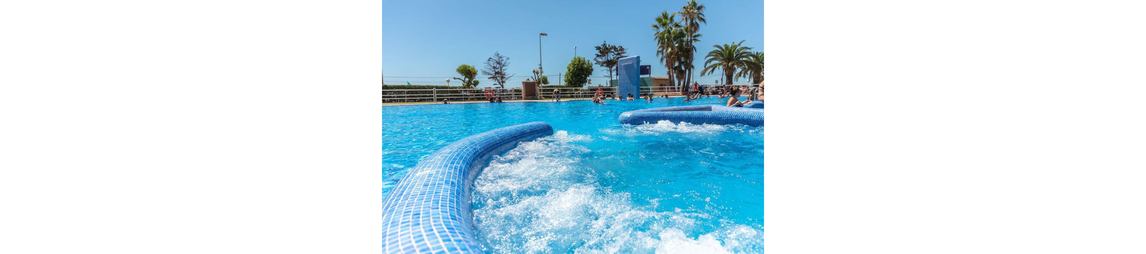 A bright blue outdoor swimming pool features bubbling water jets in the foreground. People relax and swim in the background under a clear, sunny sky. Palm trees and other greenery surround the pool area, creating a vacation resort atmosphere. The scene conveys leisure, refreshment, and summertime fun.