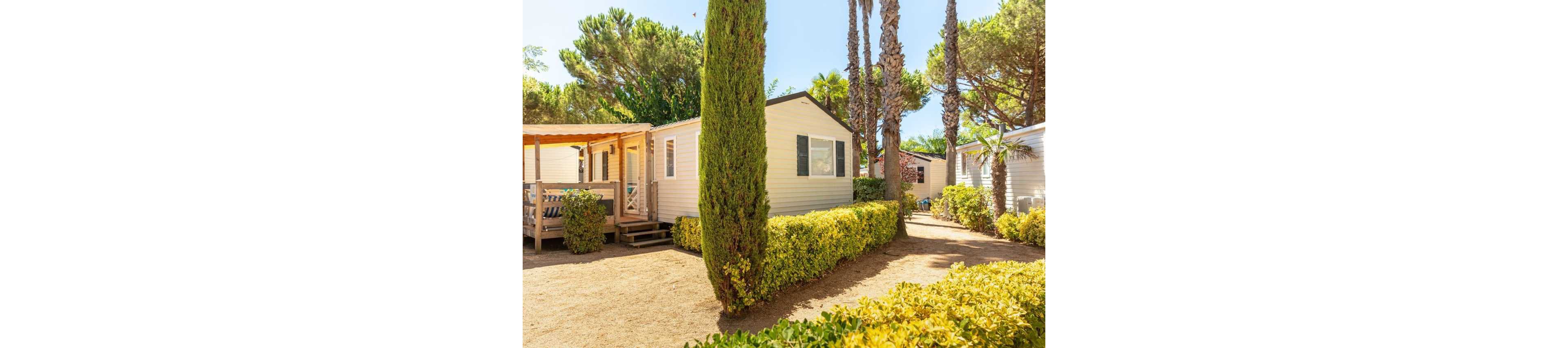 A light-colored mobile home stands in a sunny outdoor setting surrounded by tall trees and manicured hedges. A wooden deck with seating extends from the front of the home, suggesting a relaxed vacation atmosphere. The ground is covered with dry sand or gravel, and lush greenery lines the pathway between neighboring units. Bright daylight and clear skies create a warm, inviting holiday park scene.