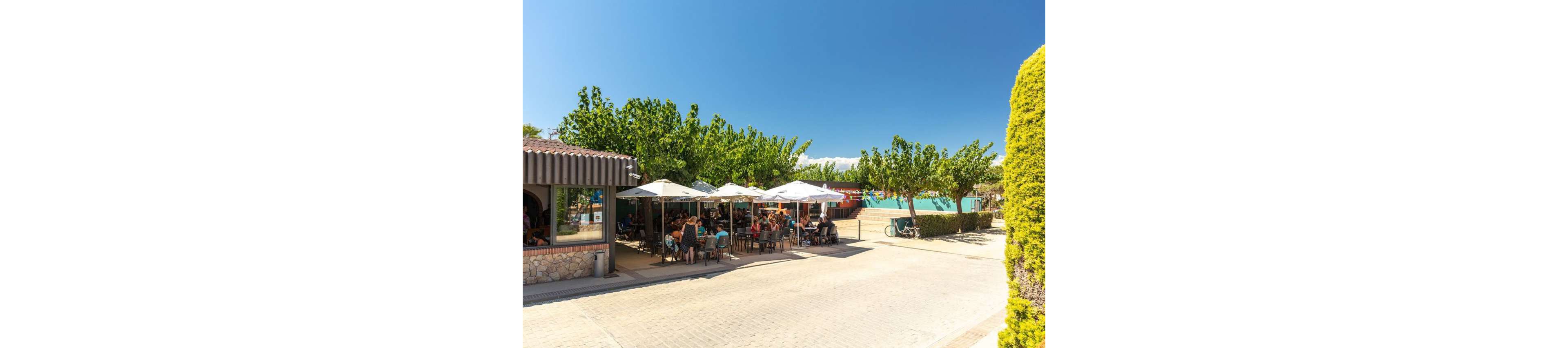 An outdoor café terrace is filled with people sitting at tables under large white umbrellas. The setting appears to be a sunny holiday destination with clear blue skies and lush green trees providing shade. A paved pedestrian area surrounds the seating, with a building on one side and colorful structures or tents in the background. Bicycles are parked nearby, suggesting a relaxed, family-friendly atmosphere.