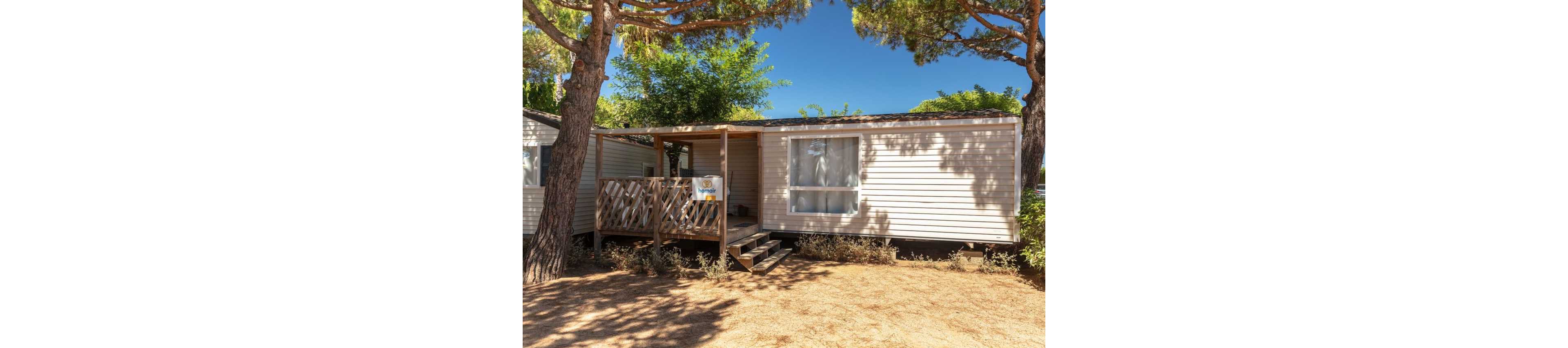 A light-colored mobile home stands beneath tall pine trees in a sunny outdoor setting. The structure features a small wooden porch with railings and steps leading up to the entrance. Large windows and a sliding door allow natural light into the interior. Dry ground and scattered shrubs surround the home, suggesting a warm, campground-like environment.