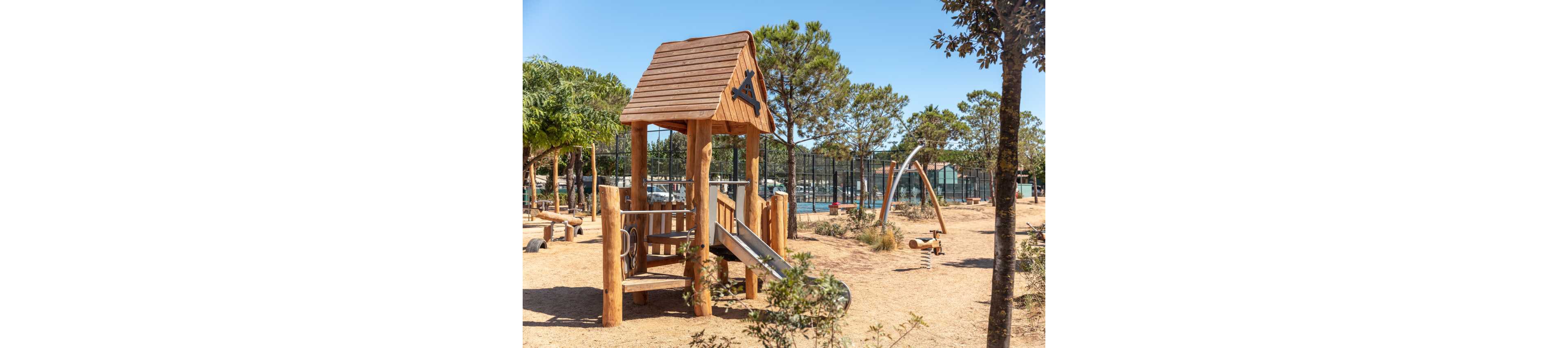 A wooden playground structure with a slide stands in a sunny outdoor park. The play area is set on dry, sandy ground surrounded by trees and shrubs. In the background, there are additional play elements and fenced sports courts. The clear blue sky and bright light create a warm, summery atmosphere.