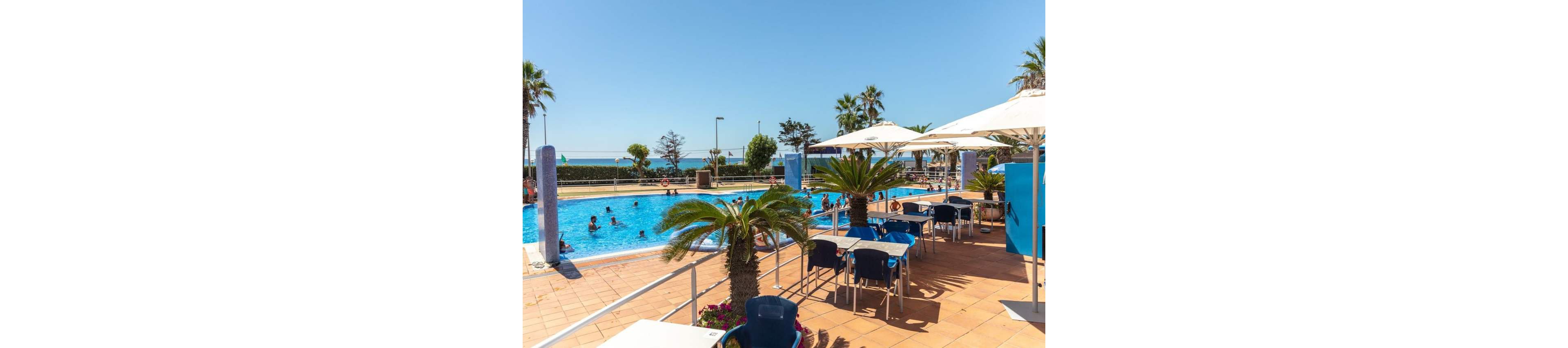 An outdoor swimming pool at a seaside resort is surrounded by sunbathers and swimmers enjoying the clear weather. In the foreground, a tiled terrace features tables, chairs, and large white umbrellas providing shade. Palm trees and potted plants add a tropical feel to the leisure area. The bright blue sky and distant ocean create a relaxed vacation atmosphere.
