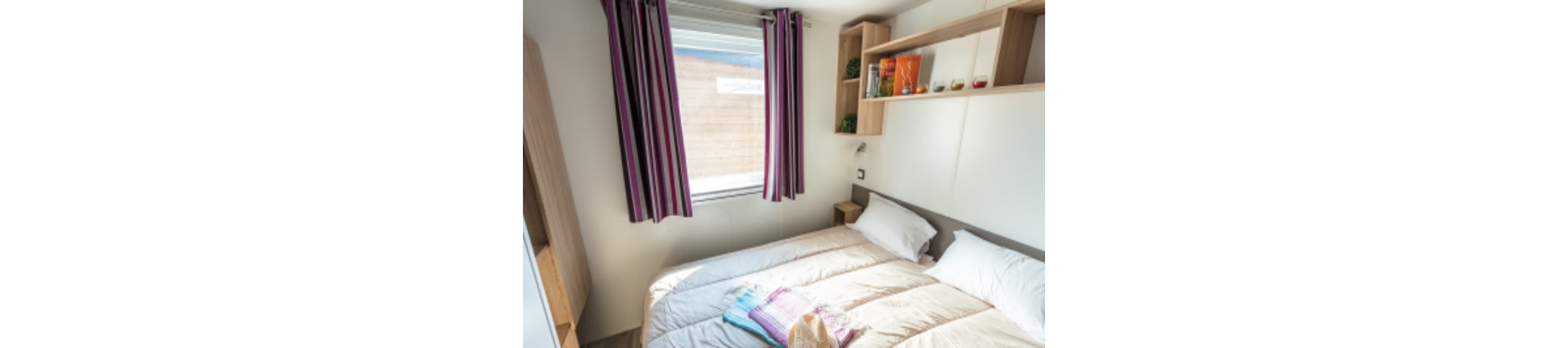 A small, neatly made double bed fills most of a compact bedroom. Sunlight enters through a window with purple and white striped curtains, brightening the neutral-toned space. Open shelves above the bed display books, plants, and small decorative items. Colorful folded towels and a straw hat rest on the bed, suggesting a cozy vacation or guest room setting.