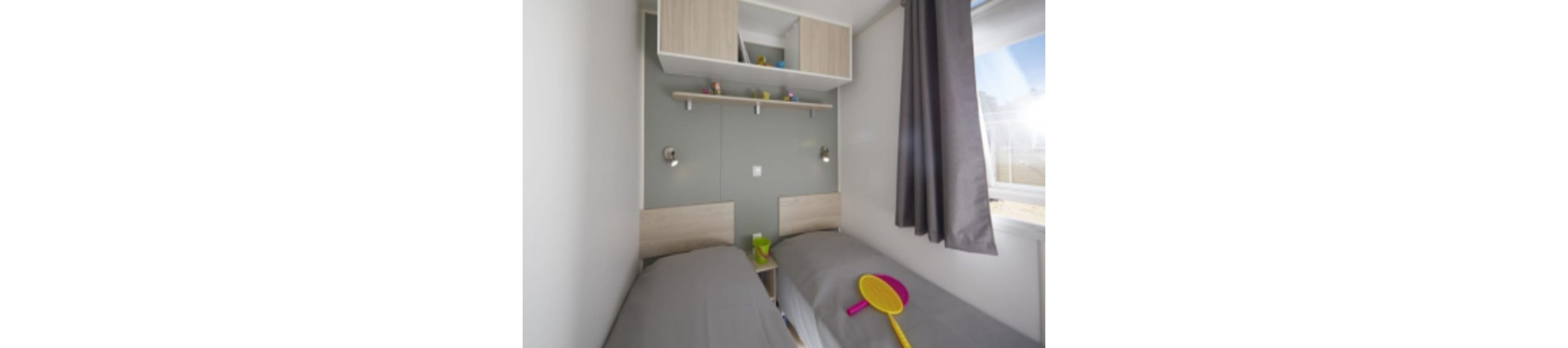 A compact bedroom features two single beds with grey duvets facing each other along the walls. Above the beds, a shelf and overhead cabinets hold small decorative items and toys. A bright yellow and pink paddle toy rests on one of the beds, adding a playful accent. Natural light enters through a window with dark curtains, giving the room a clean and modern feel.