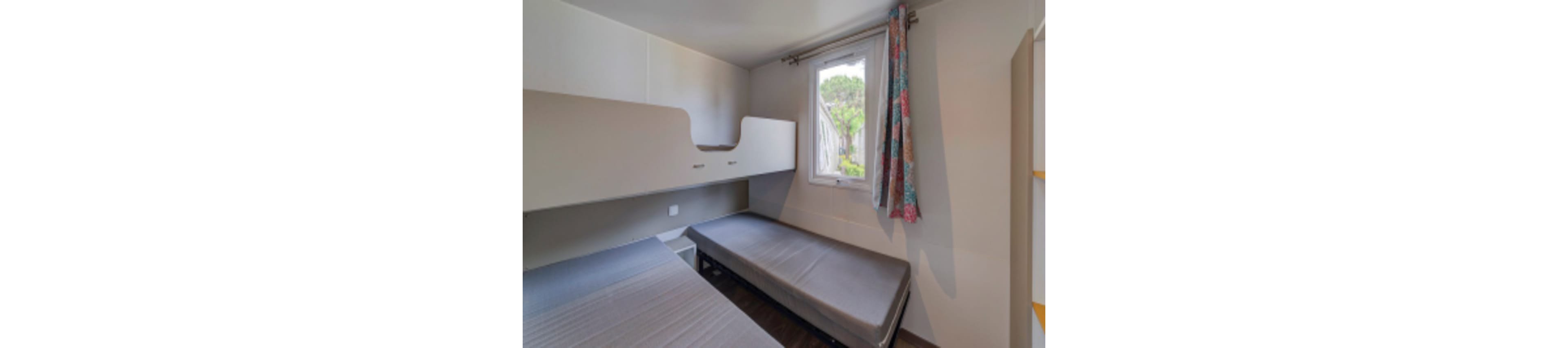 A small bedroom features two single beds arranged in an L-shape beneath built-in overhead cabinets. Natural light enters through a single window with colorful patterned curtains. The room has neutral walls and simple, modern furnishings, creating a clean and functional look. The overall space appears minimal and practical, suitable for a guest room or vacation accommodation.
