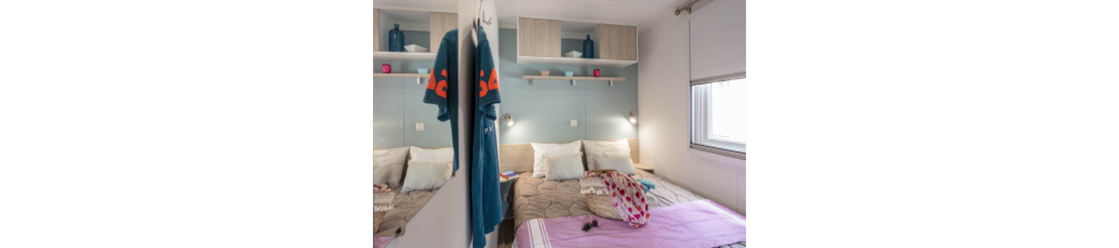 A compact modern bedroom features a neatly made double bed with neutral bedding and colorful accents. A large mirrored wardrobe door reflects the room, doubling the view of the bed and wall shelves. Decorative items such as vases, books, and small candles sit on shelves above the headboard. A window with a roller blind lets in natural light, while a towel and patterned scarf add casual, lived-in details.