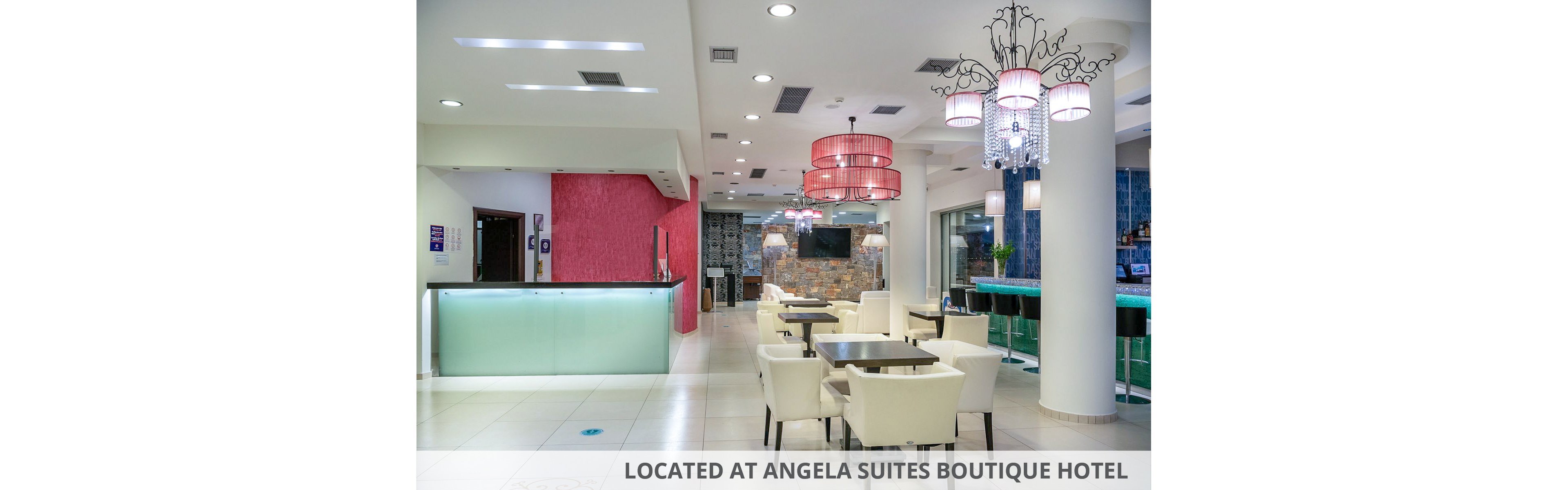 A contemporary hotel lobby features a reception desk on the left and a small seating area with white armchairs and tables in the center. In the background, a bar with high stools and blue accent tiles adds a stylish focal point. Decorative red pendant lights and a chandelier hang from the ceiling, creating a warm and inviting atmosphere. The space is brightly lit with recessed ceiling lights and finished with clean white floors and walls.