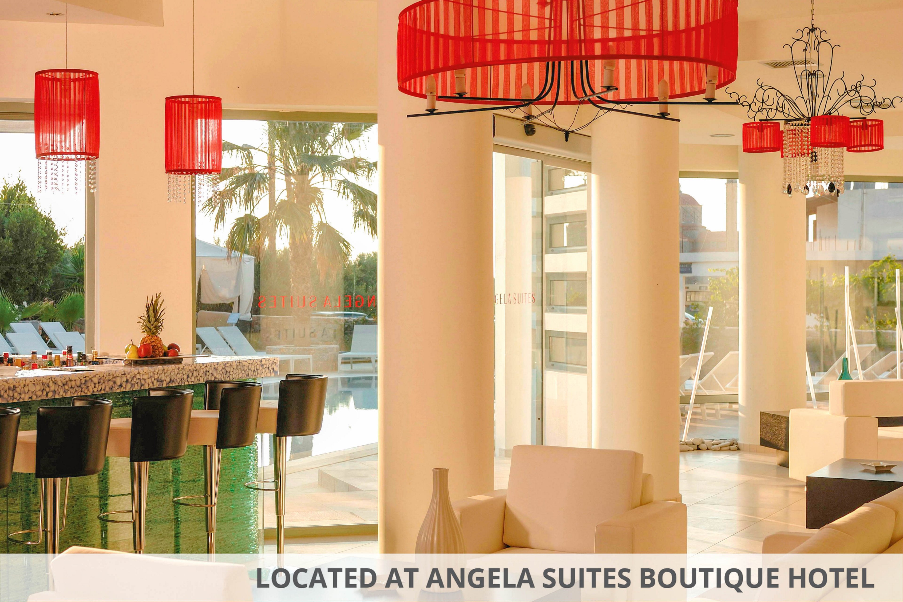 A contemporary hotel lounge bar features sleek white seating and a glossy bar counter lined with black stools. Bold red pendant lamps and chandeliers hang from the ceiling, casting warm light across the space. Large floor-to-ceiling windows reveal an outdoor pool area with palm trees and sun loungers. The overall atmosphere is bright, stylish, and inviting for guests to relax indoors while enjoying views outside.