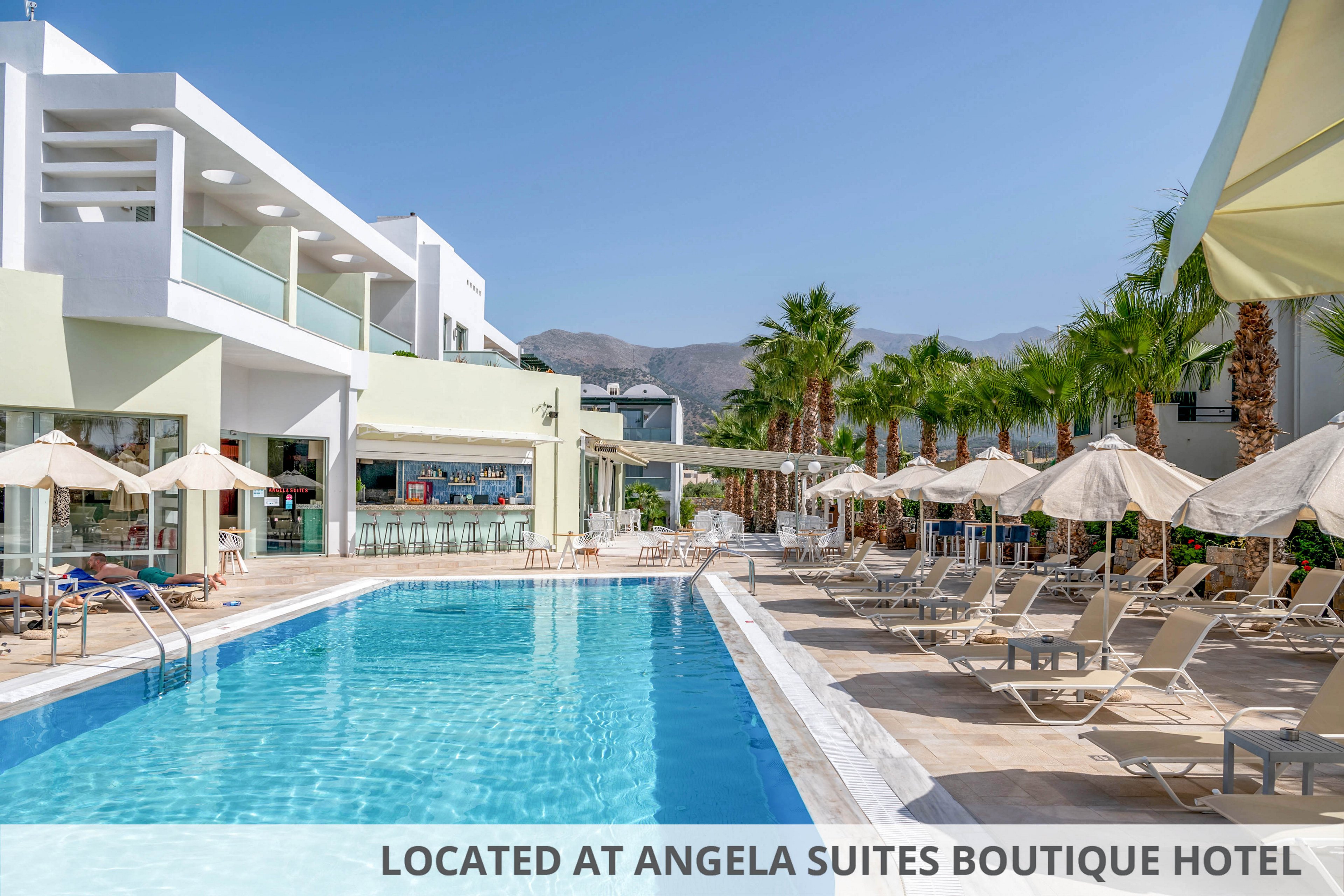 A modern resort features a long outdoor swimming pool lined with beige sun loungers and umbrellas. Contemporary white and pastel buildings frame the pool area, with a poolside bar visible in the background. Tall palm trees and distant mountains create a relaxed vacation atmosphere under a clear blue sky. The scene is bright and inviting, suggesting a sunny day at a holiday destination.