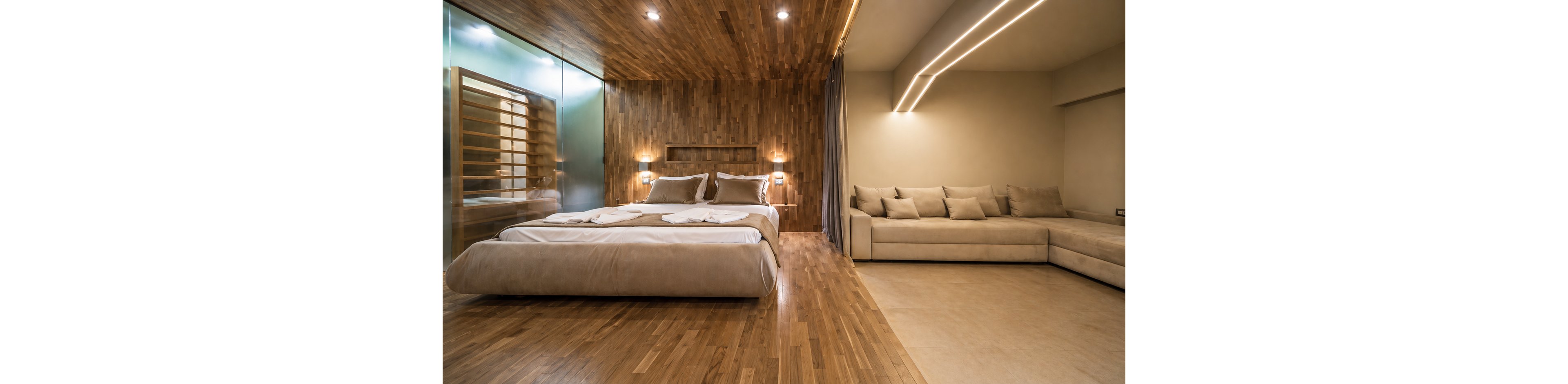 A spacious bedroom features a large bed with beige linens and a matching sofa, set against wood-paneled walls and ceiling. The room is illuminated by recessed lighting and sleek linear fixtures, creating a warm and inviting atmosphere. Glass partition and minimalist decor enhance the contemporary style, with no visible text or numbers present.