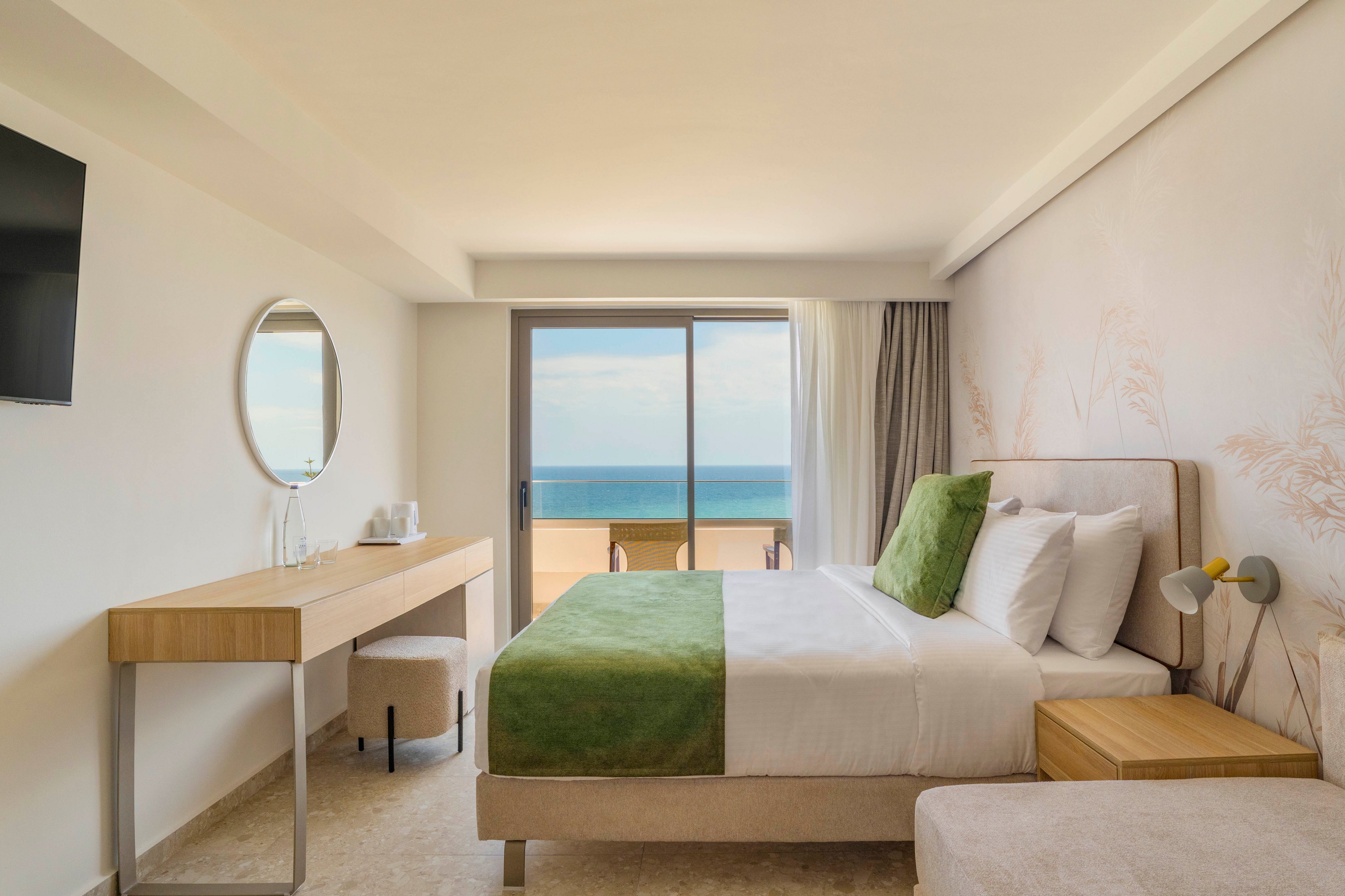 A contemporary hotel bedroom features a neatly made bed with white linens and green accents. A large sliding glass door opens to a balcony overlooking a calm ocean horizon. The room includes a minimalist wooden desk with a round mirror, a wall-mounted television, and soft neutral tones. Natural light fills the space, creating a bright and relaxing atmosphere ideal for a seaside stay.