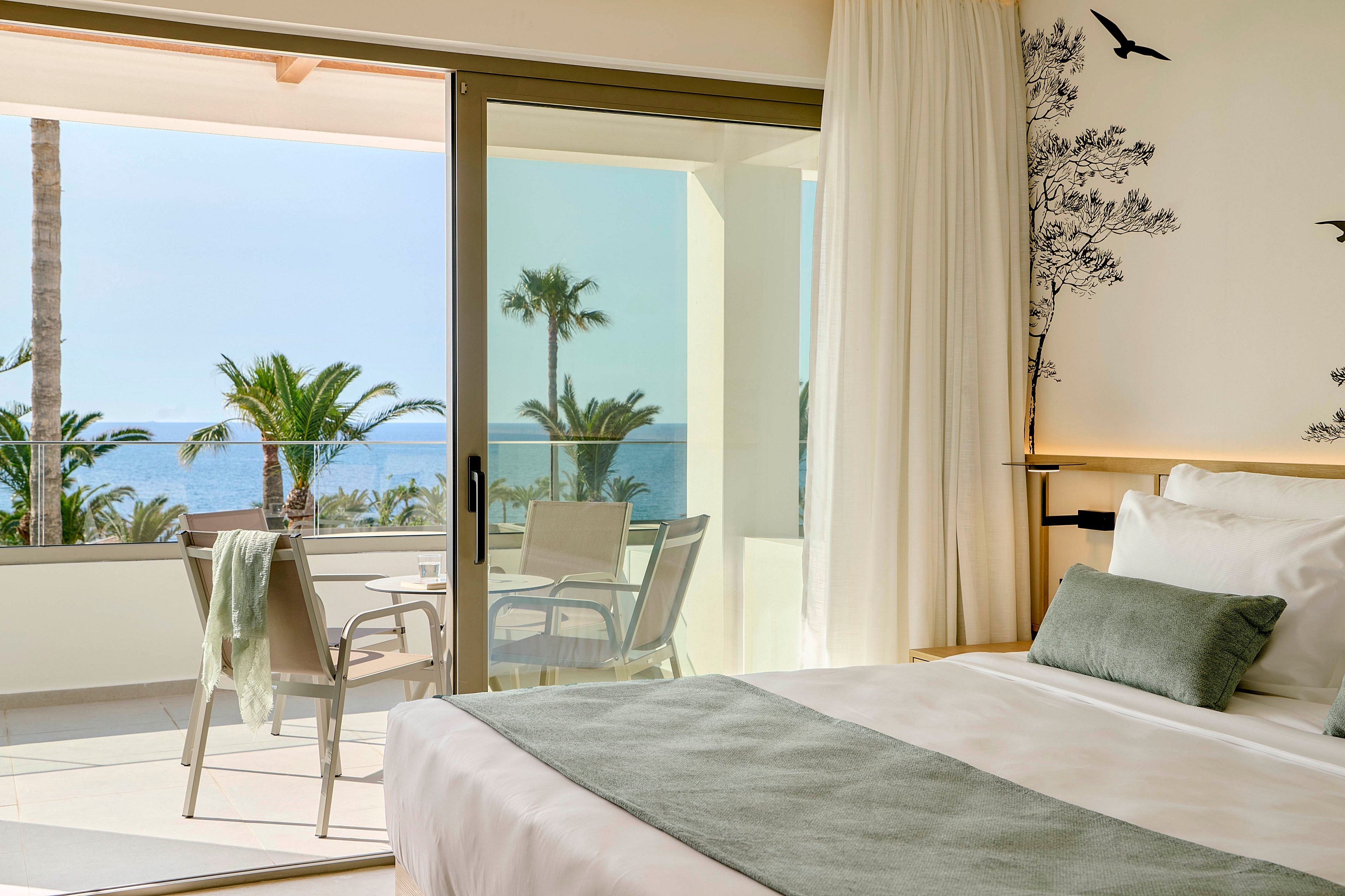A contemporary hotel bedroom features a neatly made bed with soft white linens and a pale green throw. Large sliding glass doors open onto a private balcony furnished with chairs and a table. Beyond the balcony, palm trees frame a clear view of the ocean under a bright sky. Soft natural light and minimalist decor create a calm, relaxing atmosphere ideal for a coastal getaway.