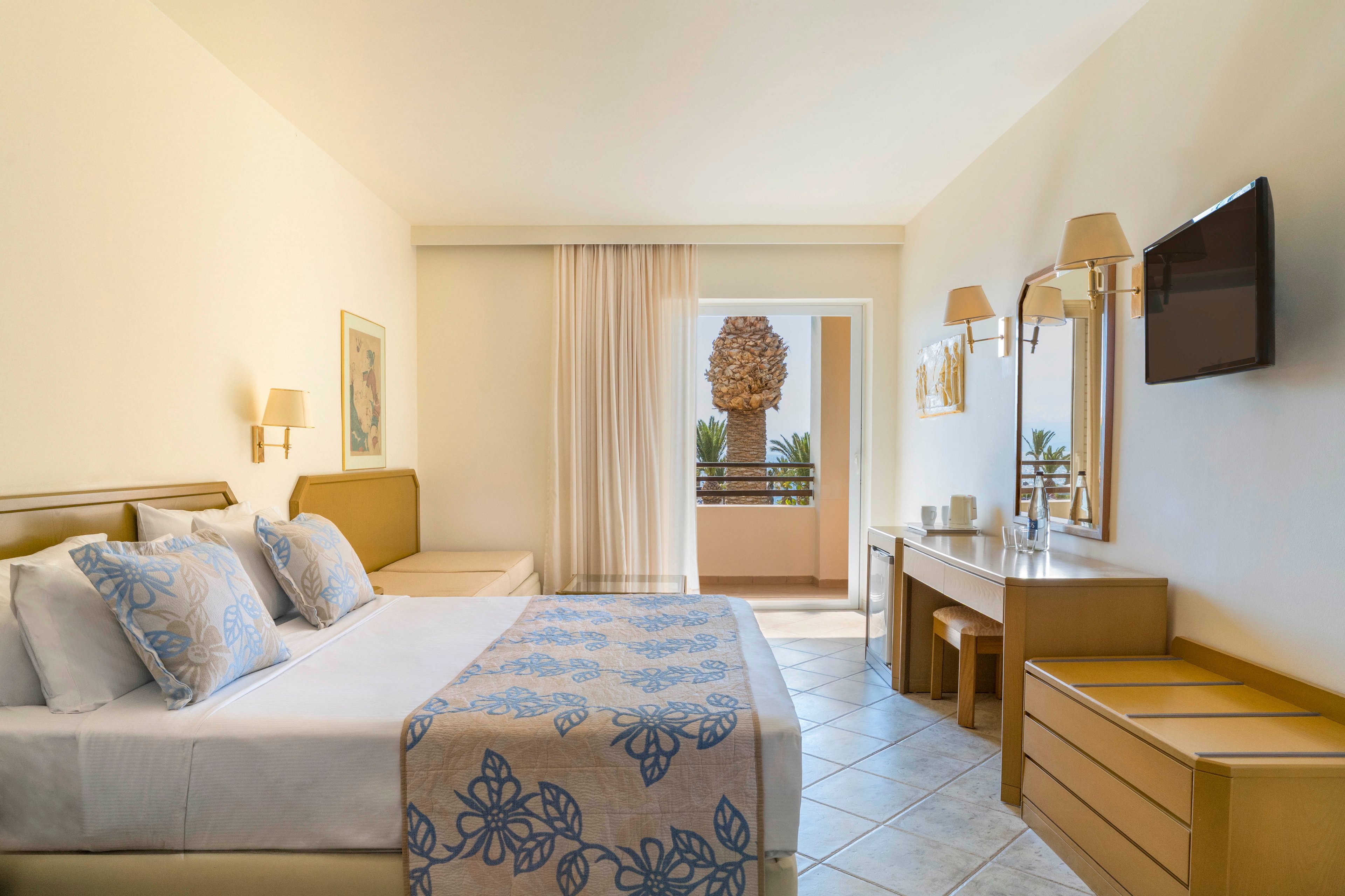 A neatly made double bed with patterned blue bedding sits in a bright, modern hotel room. The space features light wood furniture, including a desk with a mirror, wall lamps, and a mounted flat-screen television. Sheer curtains partially cover a glass door that opens to a balcony with potted plants and an outdoor sculpture. Soft neutral tones and natural light create a calm, inviting atmosphere suitable for vacation or business travel.