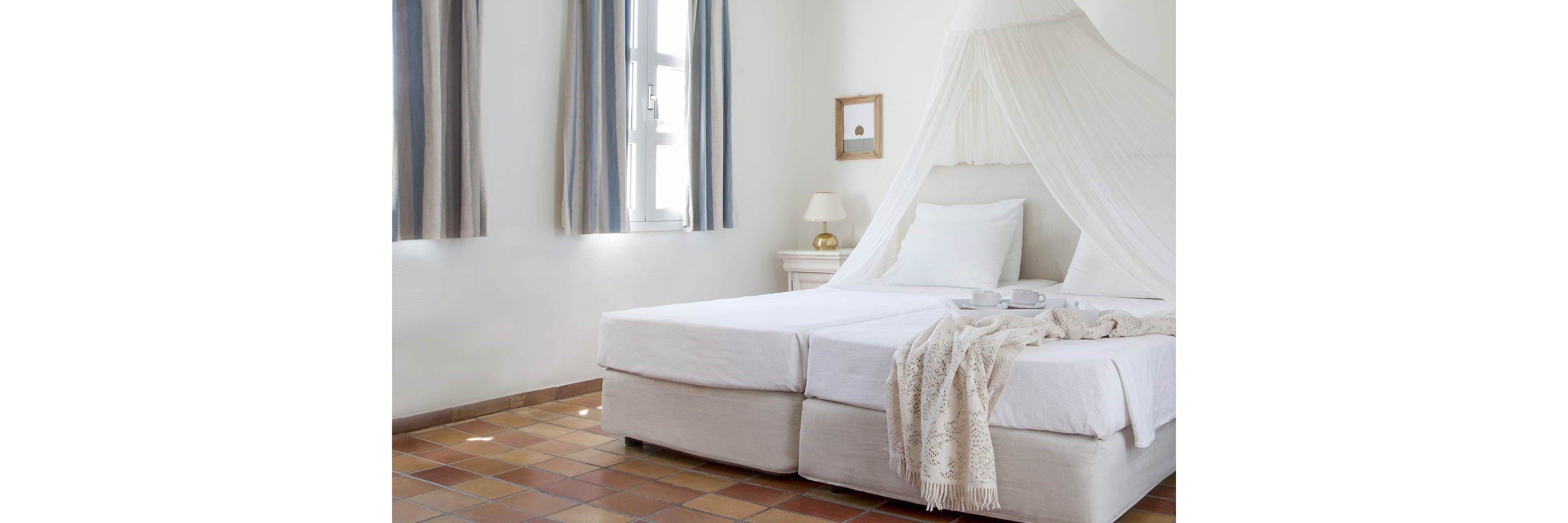 A light-filled bedroom features a white double bed with a sheer canopy and crisp linens. The space is decorated in a minimalist style with neutral tones, terracotta tile flooring, and simple wall art. A small bedside table with a lamp and a tray holding cups adds a cozy, welcoming touch. Sunlight streams through the windows with soft curtains, enhancing the airy and serene atmosphere.