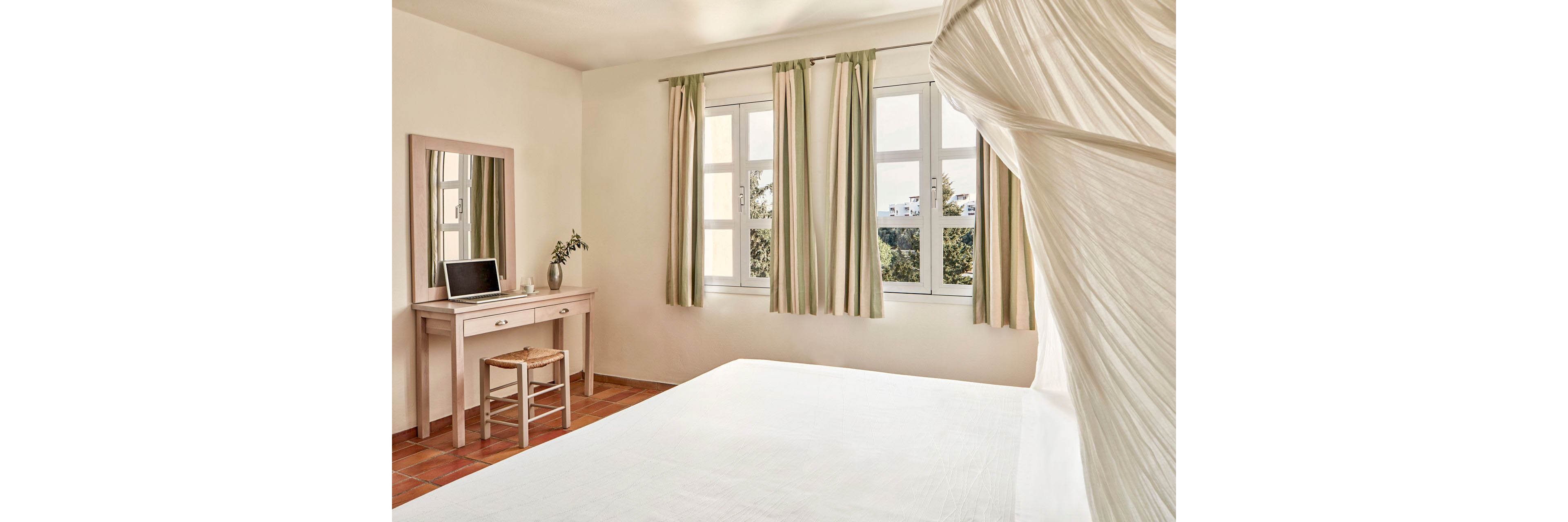 A sunlit minimalist bedroom features a large white bed in the foreground and a simple wooden desk with a mirror and stool along the wall. A laptop sits on the desk beside a small vase with greenery, adding a modern touch to the serene space. Multiple tall windows with light green curtains let in natural light and reveal trees and distant buildings outside. Warm terracotta floor tiles and soft neutral tones create a calm, airy atmosphere.