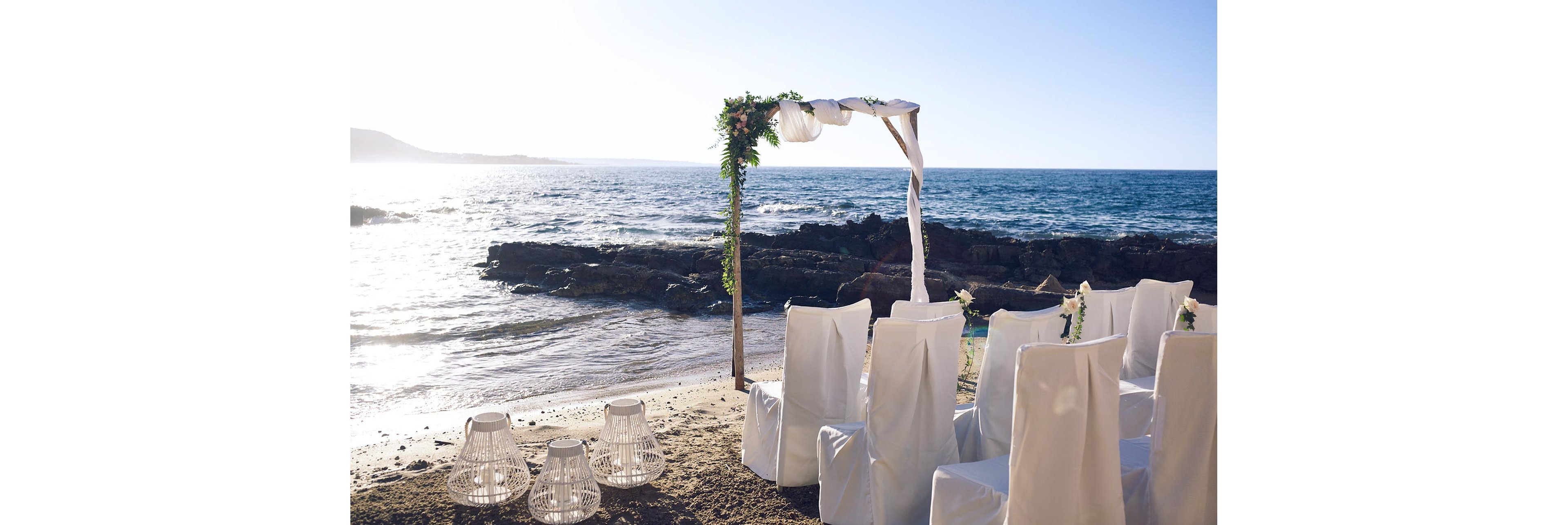 A wedding ceremony setup is arranged on a sandy beach facing the ocean. White-covered chairs form neat rows beneath a simple wooden arch decorated with greenery and white fabric. Decorative white lanterns sit on the sand in the foreground, adding a romantic touch. The calm sea, rocky shoreline, and low sun create a serene, golden-hour atmosphere.